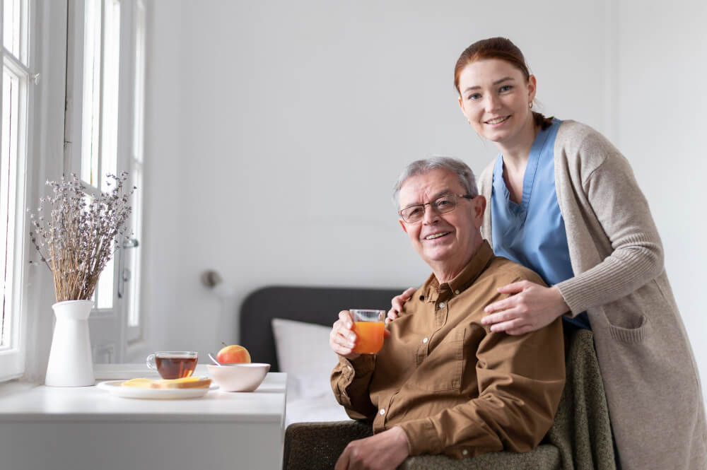 A caregiver smiling with an elderly man enjoying a drink in a cozy room.