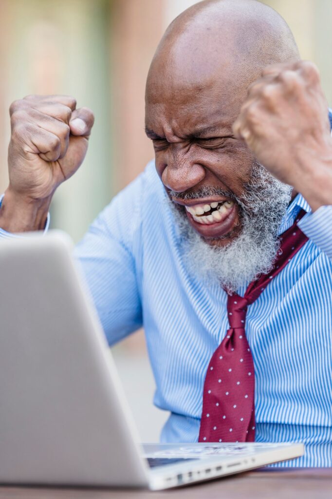 Blog Pix Clutter - For My Parents Care Elderly man with a gray beard in a blue shirt and red tie clenches fists in while looking at a laptop